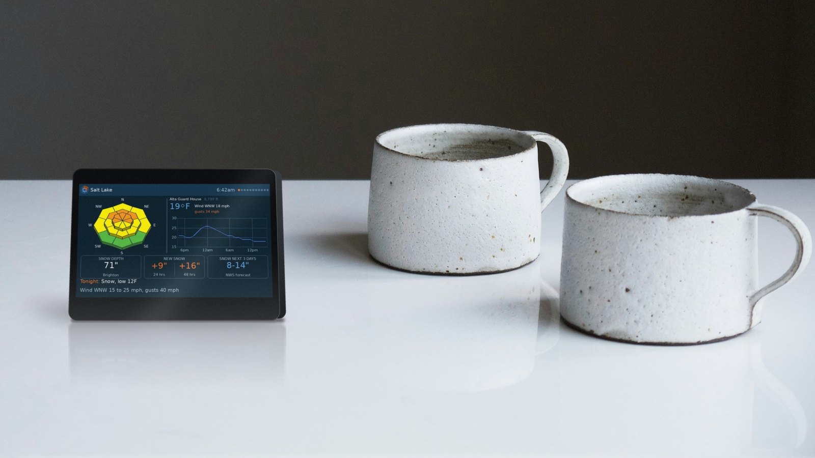 PowDash on its stand on a wooden kitchen counter next to a coffee mug, about 4 inches wide and 3 inches tall